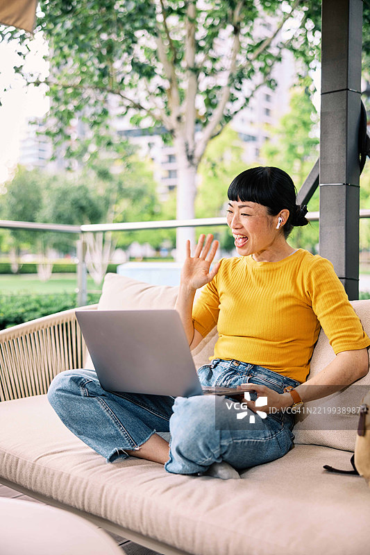 友好的日本女性坐在阳台上，膝上放着笔记本电脑，正在视频通话中挥手。图片素材