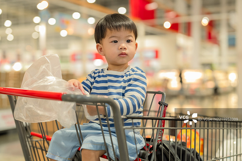 快乐的小男孩坐在明亮超市的购物车里，开心地观察周围，笑声不断。他穿着休闲服，散发着天真和快乐，带来温暖和日常家庭的感觉。图片素材