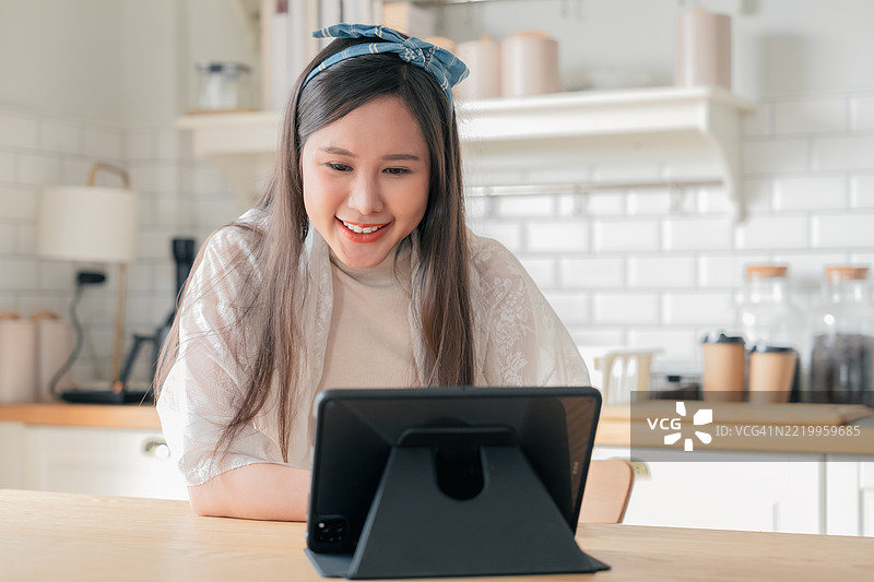一位年轻美丽的亚洲女性在家中的厨房里独自享受吃面条的乐趣。她在平板电脑上观看电影，营造出一个温馨宁静的家庭时刻，反映出现代生活方式和舒适食品文化。图片素材