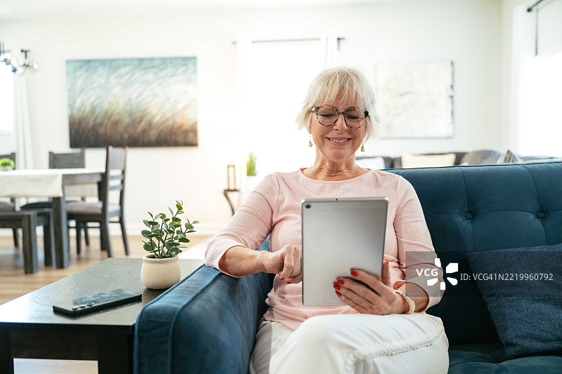 一位老年女性在客厅里微笑着喝咖啡，同时使用她的平板电脑。图片素材