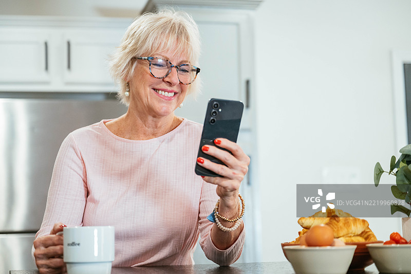 一位老年女性在明亮的厨房里微笑着喝咖啡，同时使用她的智能手机。图片素材