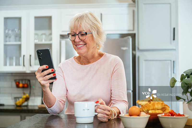 一位老年女性在明亮的厨房里微笑着喝咖啡，同时使用她的智能手机。图片素材
