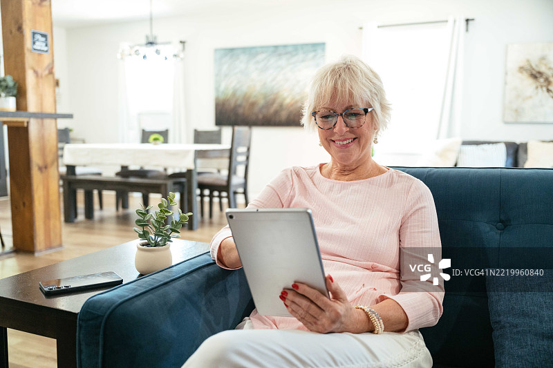 一位老年女性在客厅里微笑着喝咖啡，同时使用她的平板电脑。图片素材