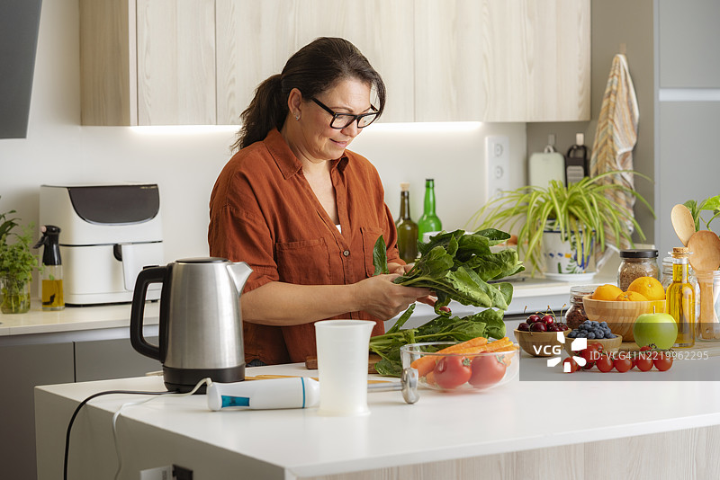 女性在现代厨房准备蔬菜餐。使用家用电器烹饪健康食品。厨房岛上摆放着水果和蔬菜。图片素材