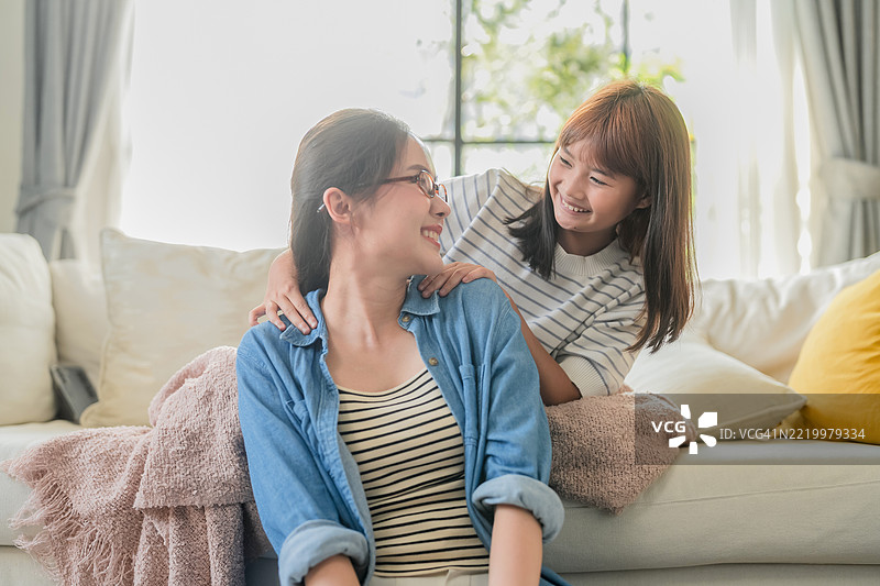 一个充满爱的亚洲女儿从后面拥抱着微笑的母亲，同时轻柔地按摩她的肩膀，坐在温馨的客厅里。她们快乐的笑声和温暖的拥抱反映了深厚的情感纽带、家庭的亲情以及跨代爱的美好。图片素材