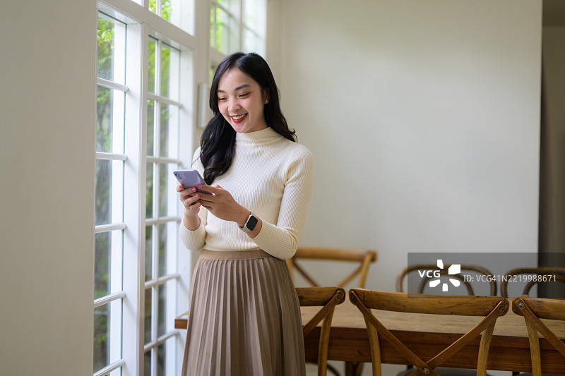 愉快的亚洲女性在窗边使用智能手机，阳光照亮了她在舒适家居环境中的放松姿态图片素材