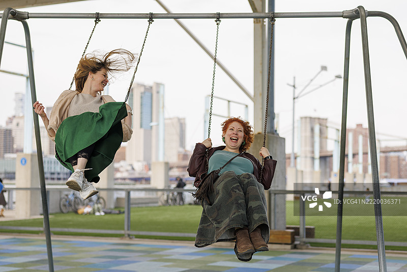四十多岁的朋友们在游乐场的秋千上尽情享受。布鲁克林的天际线为这个无忧无虑的时刻创造了城市背景。图片素材