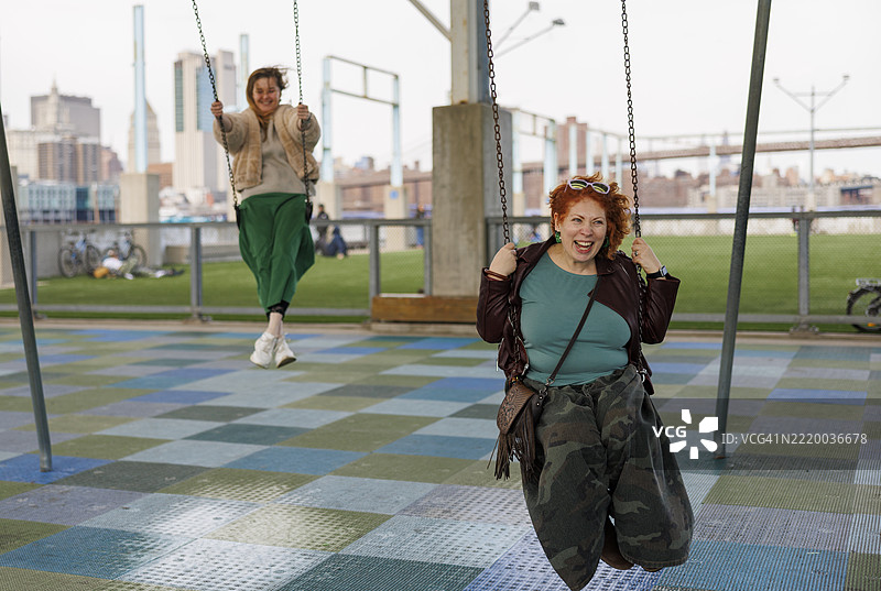 成年人在秋千上享受游乐场的乐趣。布鲁克林公园背景是曼哈顿天际线。图片素材