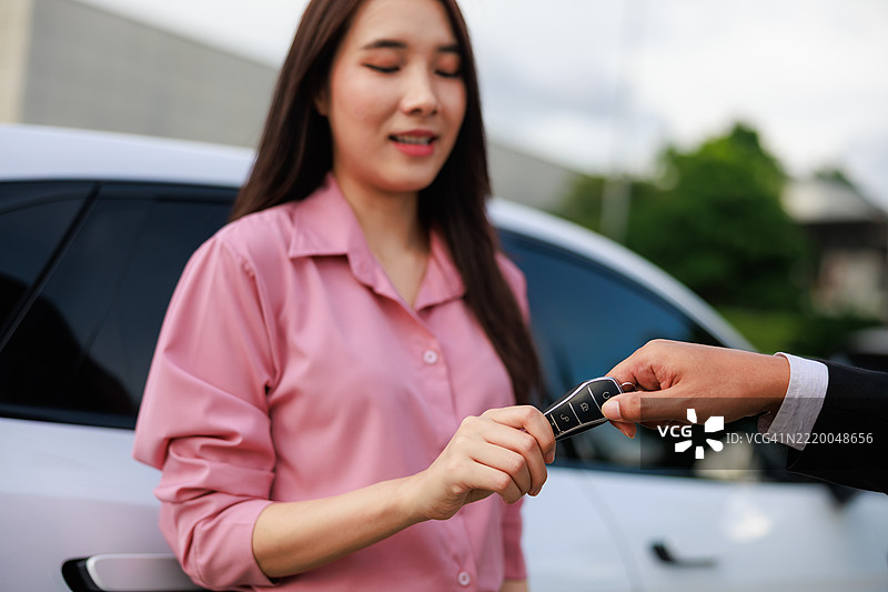 一位亚裔女性在明亮现代的汽车经销商展厅内，从销售人员手中接过车钥匙。这张照片捕捉了购买新车的瞬间，突显了客户满意度、生活方式的追求以及汽车销售的重要性。图片素材