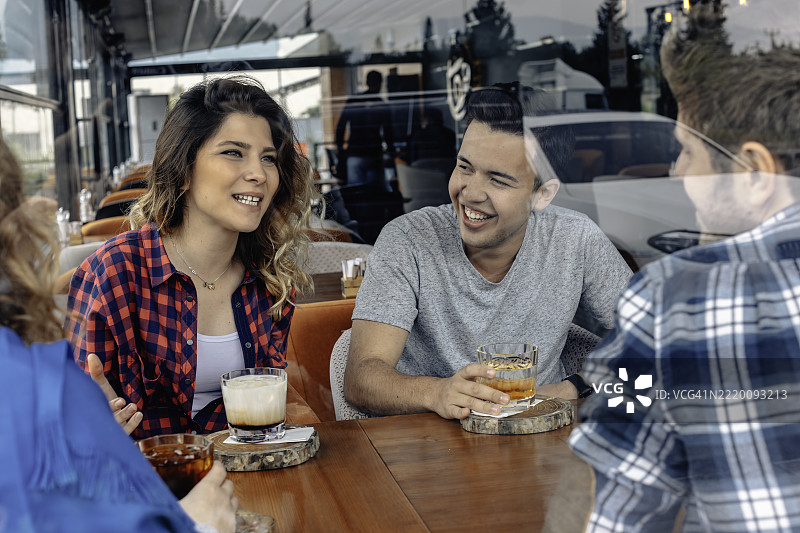 年轻人坐在咖啡桌旁，透过玻璃享受咖啡鸡尾酒的社交时光图片素材