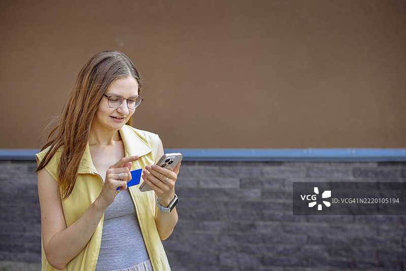 戴眼镜的年轻女子自信地拿着银行卡和智能手机，展现她的独立。图片素材