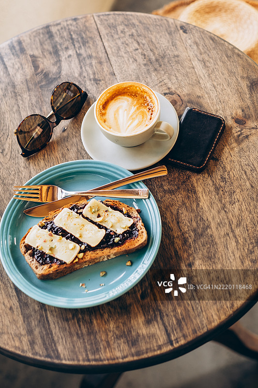 户外咖啡馆风格的早餐，有涂抹果酱的烤面包和布里奶酪片，旁边还有卡布奇诺咖啡、太阳镜、钱包和草帽，摆放在乡村风格的圆桌上。图片素材