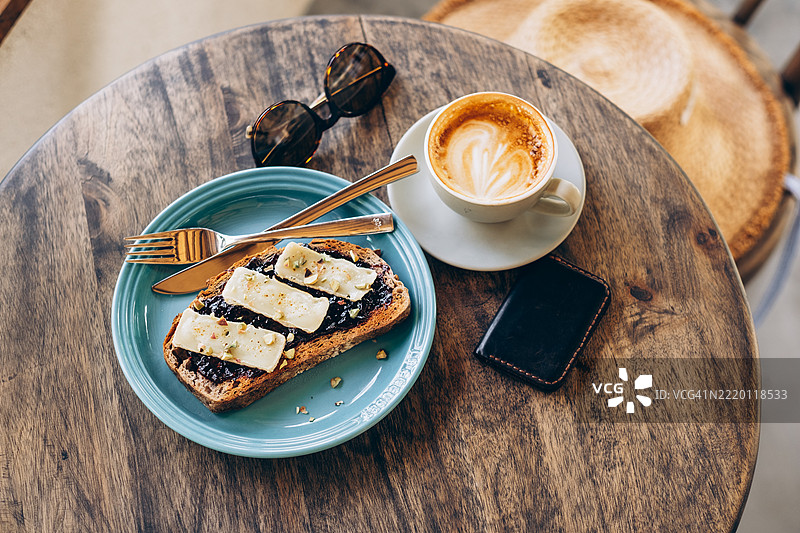 咖啡和涂有融化布里奶酪和果酱的吐司，摆放在粗犷的木桌上，旁边有太阳镜、草帽和钱包。图片素材