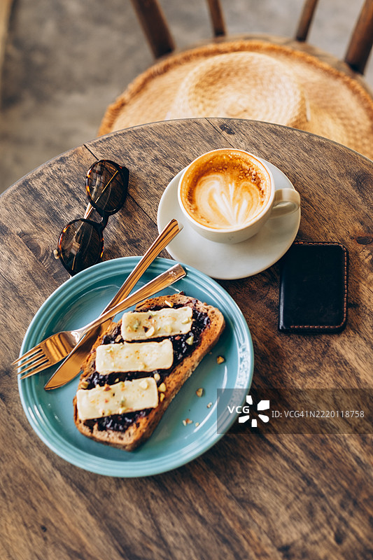 咖啡馆早午餐桌俯视图，包括卡布奇诺、蓝盘上的布里奶酪吐司、钱包、太阳镜以及椅子上的草帽。图片素材