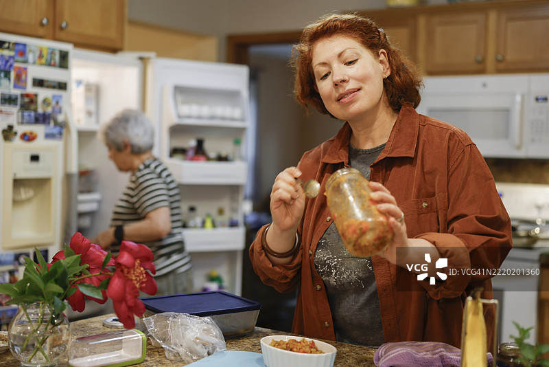 腌制蔬菜。中年女性在宾夕法尼亚州乡村厨房检查自制莎莎酱的罐子，而祖母则在忙碌。图片素材