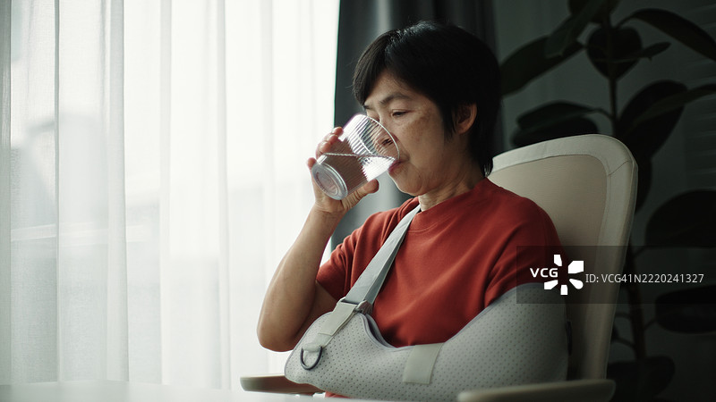 戴着手臂石膏的成熟的女性在喝水。图片素材