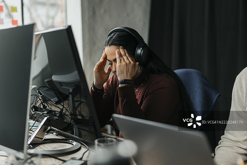疲惫的女性程序员坐在科技初创公司的办公桌前，捂着太阳穴。图片素材