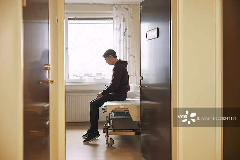 悲伤的少年坐在诊所的检查台上，从门口看过去。图片素材
