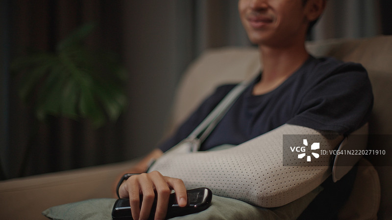 晚上，手臂打着石膏的男人在沙发上放松地看电影。图片素材