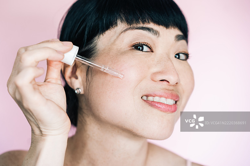 亚洲女性使用滴管涂抹面部精华。深青色背景与护肤程序形成对比，突显美丽、青春和健康的肌肤护理。图片素材