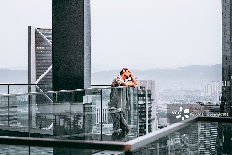 中年白人男性站在现代城市上方的玻璃阳台上。城市景观中有高楼大厦。图片素材