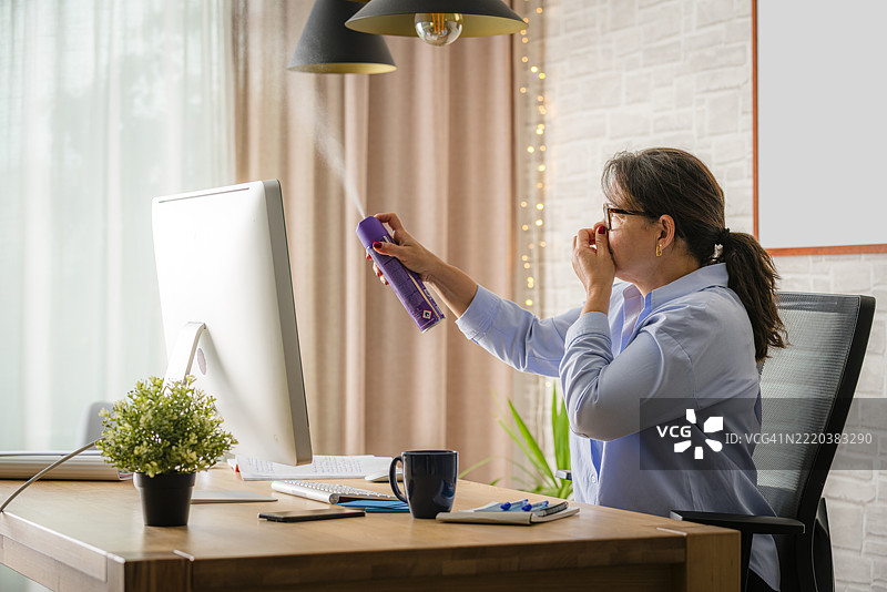 女性使用空气清新剂去除办公室或家中的异味，闻到令人厌恶的臭味，商务女性在桌子上工作，使用电脑。图片素材