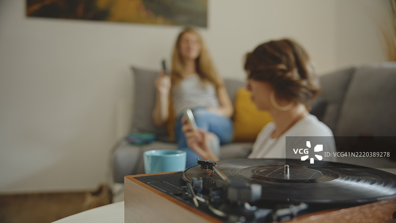 两个白人女性朋友在舒适现代的客厅里听黑胶唱片，享受咖啡。图片素材