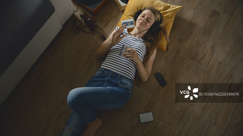 年轻女性坐在地板上，戴着复古耳机，听着音乐，旁边有一盘卡带。图片素材