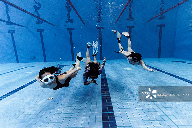 三位穿着泳衣和脚蹼的女性在游泳池中潜水，练习同步动作和自由潜水技巧。图片素材