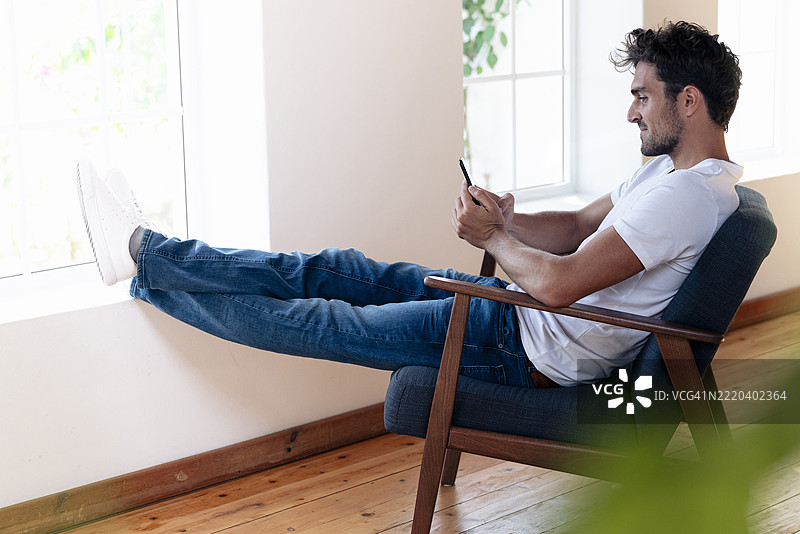 男子坐在家中窗边，双腿交叉，使用手机。图片素材