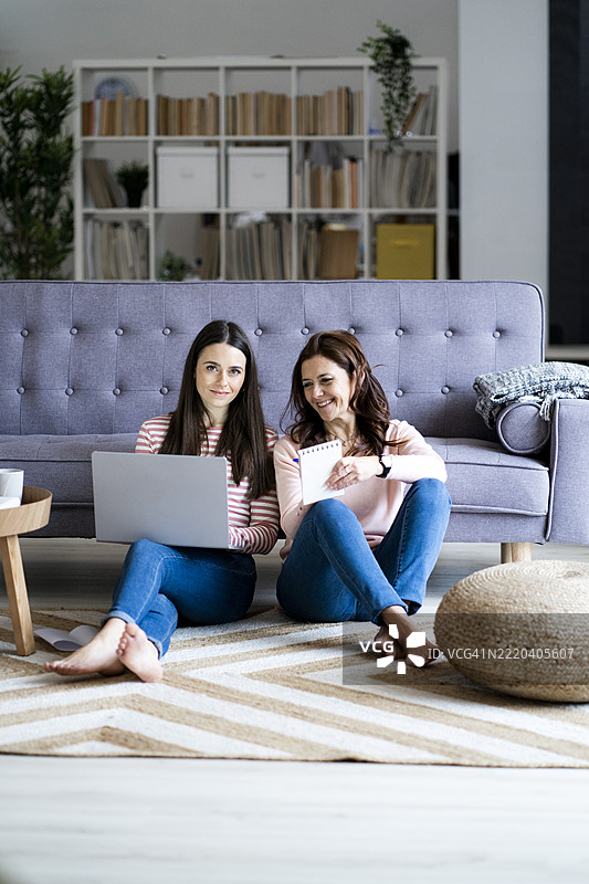 微笑的年轻女性坐在家中地板上，旁边是她的母亲，手里拿着笔记本电脑。图片素材