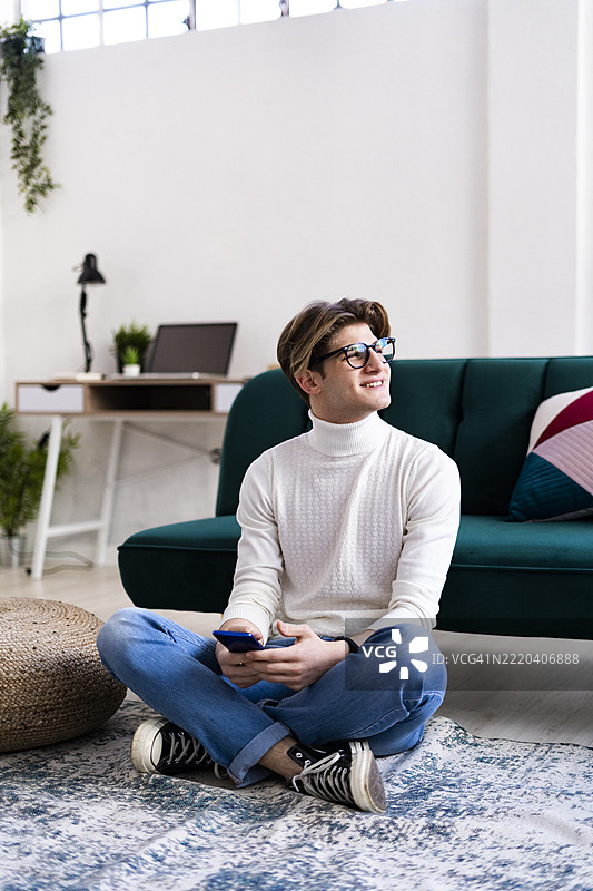 微笑的年轻男子坐在客厅的地毯上，手持智能手机，目光投向一旁。图片素材