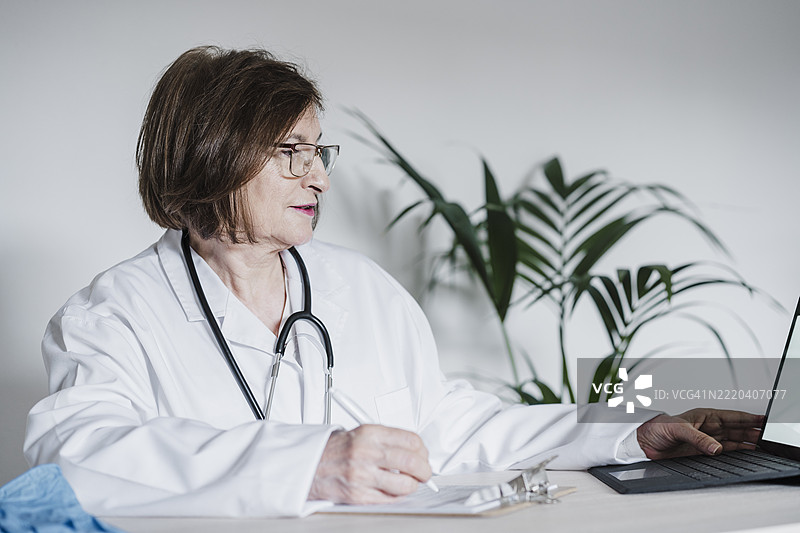 女医生在医疗诊所的桌子上使用数字平板电脑，同时在夹板上写报告。图片素材