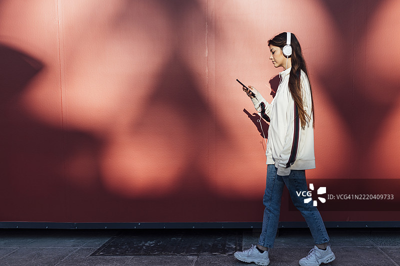 一名少女在红色墙壁旁边走路时使用智能手机听音乐图片素材