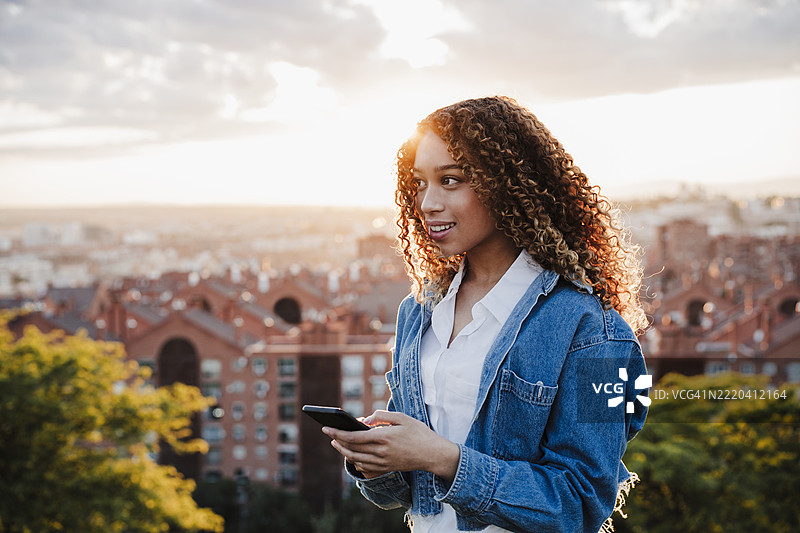 西班牙裔女性在日落时望向远方，手中拿着智能手机图片素材