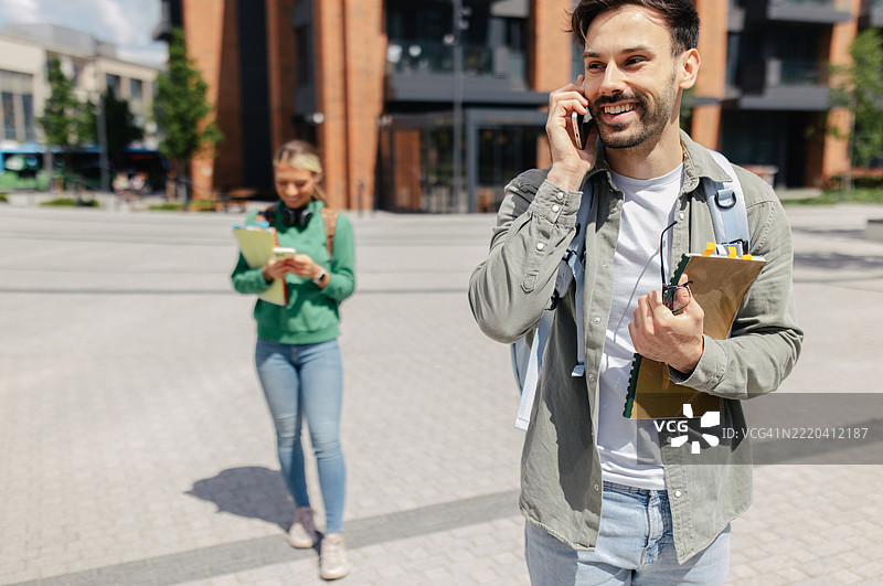 一名男子在打电话，面带微笑，背景中有一名学生。图片素材