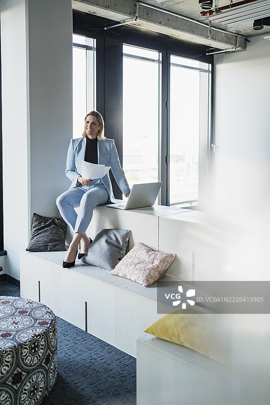 微笑的女性企业家坐在办公室的家具上，手里拿着文件和笔记本电脑，目光投向一旁。图片素材