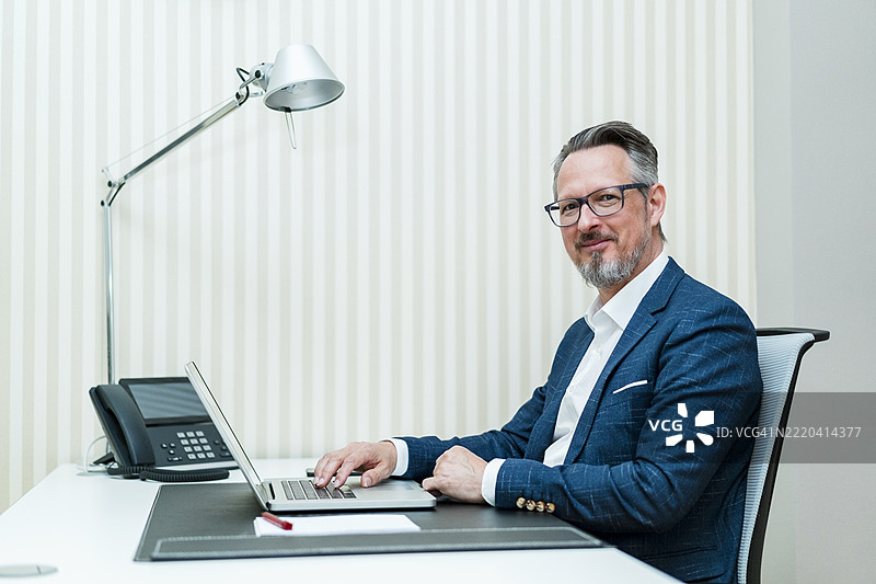 成熟的商人在办公室的桌子旁坐着，使用笔记本电脑图片素材