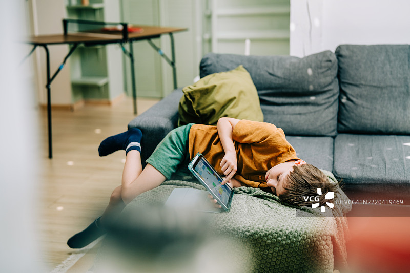 无聊的男孩躺在家里的沙发上使用平板电脑。图片素材