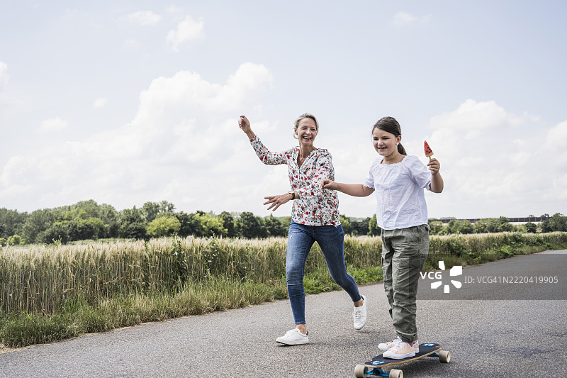 微笑的女人在滑板上站着的女孩旁边跑过图片素材