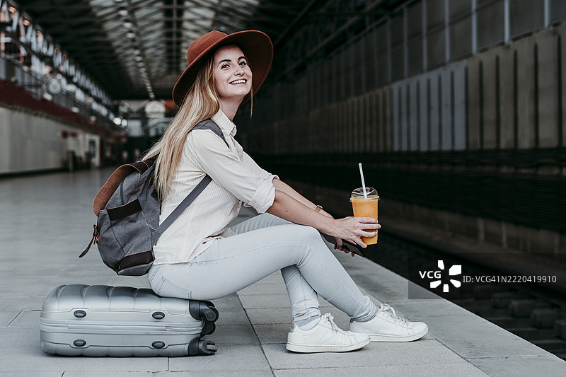 微笑的年轻女性乘客坐在行李上，手里拿着果汁，站在火车站月台上。图片素材