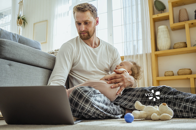 商人在家中使用笔记本电脑，女儿安静地睡在他的腿上。图片素材
