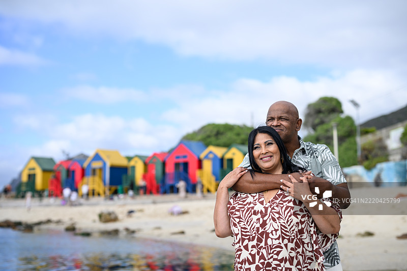 在穆伊岑堡海滩，一对幸福的情侣相拥而立，背景是五彩斑斓的小屋和蓝天。图片素材