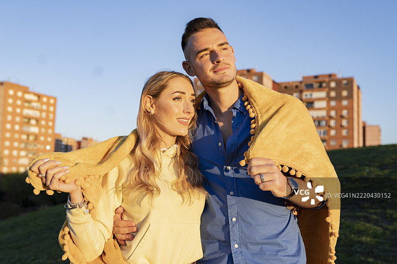 年轻情侣裹着毯子站在日落时的山丘上图片素材