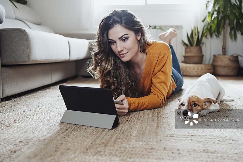 女人在家中躺在地板上使用平板电脑，身旁有一只杰克拉塞尔犬。图片素材