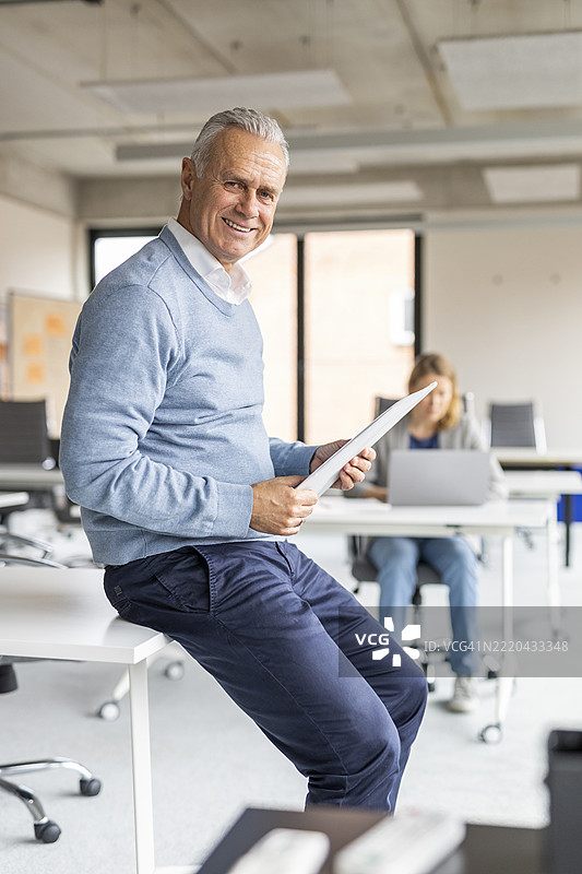 微笑的老年商人坐在办公室的桌子上，手里拿着文件夹。图片素材