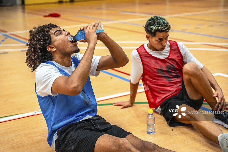 两名篮球运动员坐在球场上，休息并在激烈的训练后喝能量饮料。图片素材