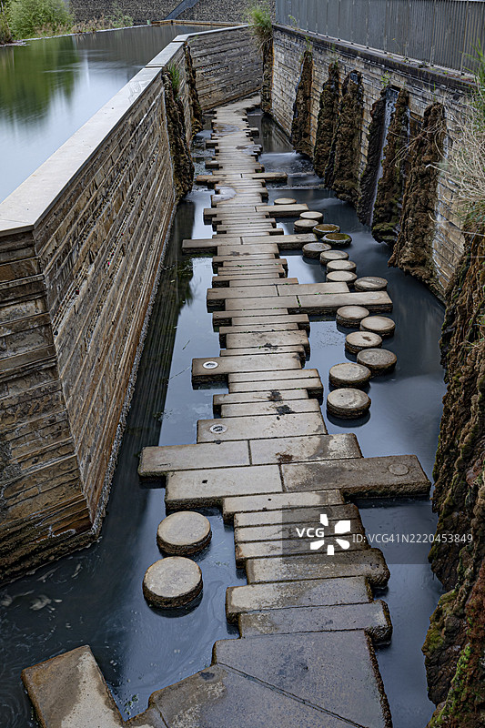 一条石头小路穿过一条狭窄的水道，旁边是一面墙，位于德国萨尔州的兰茨维勒-雷登水景花园。图片素材