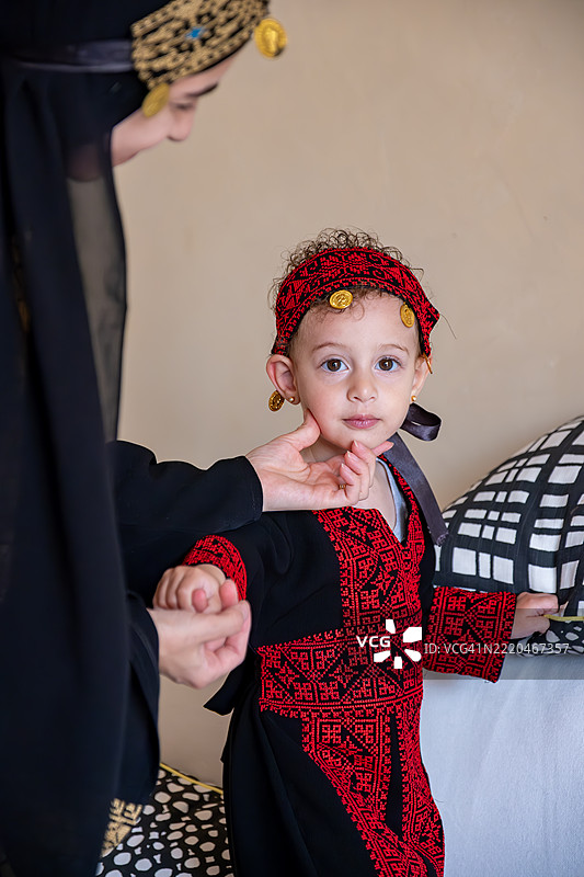 一个年轻的孩子穿着传统的红黑相间的巴勒斯坦刺绣裙子，配有相应的头带和装饰性硬币，站在一位穿着类似传统服装的女性旁边，背景是一个现代家庭环境。图片素材
