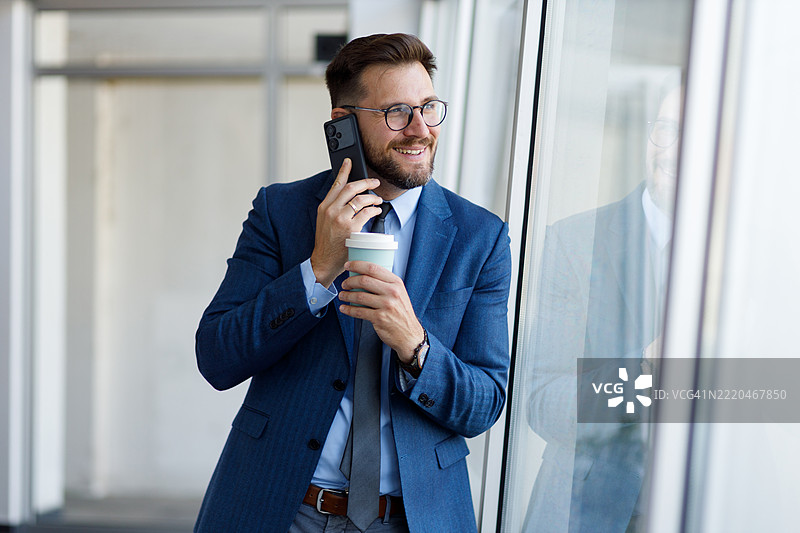 专业商务人士通过智能手机沟通，边喝咖啡边凝视办公室走廊的窗户。图片素材